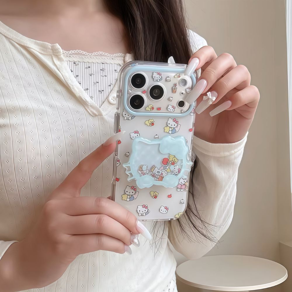 A person with long white almond-shaped nails holding a clear iPhone case decorated with a Hello Kitty and little kawaii pattern. The case features a light blue camera protector and a matching blue Hello Kitty shaker pop socket with character charms inside.