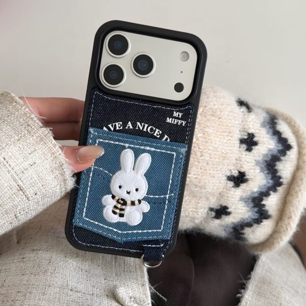 An image of a hand wearing a light-colored tweed jacket sleeve holding a phone case, as seen in. The black-framed phone case has a dark denim back featuring an embroidered Miffy bunny wearing a scarf within a lighter denim pocket, white text reading "HAVE A NICE DAY," and "MY MIFFY" at the top right. A silver D-ring is attached to a denim strap at the bottom left of the case. The phone has a silver camera island with three large lenses. A person's fingers with manicured nails are visible. The background includes a beige and dark grey patterned knit blanket.